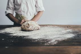Photographie d'une boulangère ou un boulanger devant une planche où une pâte est déposée sur la surface enfarinée.