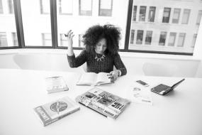 Photographie en noir et blanc d'une personne assise à une table avec plusieurs livres devant elle. Un ordinateur portable et un téléphone intelligent repose également sur la table.