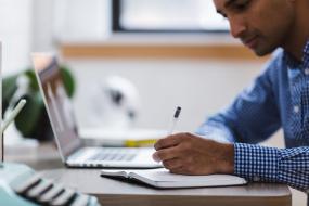 Une personne écrit sur une table, un ordinateur portable est placé à sa gauche.