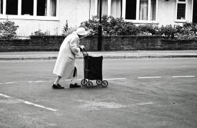 Photographie en noir et blanc d'une femme âgée poussant un chariot portatif dans un stationnement vide.