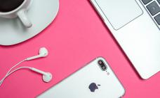 Sur une table rose, on voit partiellement un café, un ordinateur portable, un téléphone intelligent et ses écouteurs.