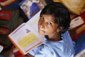 Un enfant assis par terre avec un livre, regard l'objectif de la caméra.