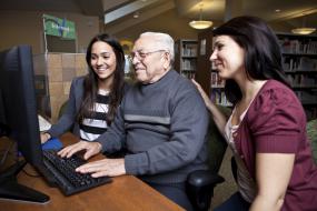 Deux jeunes encadrent une personne âgée utilisant un ordinateur.