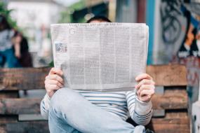 Photographie d'une personne assise sur un banc public tenant un journal. Le journal cache complètement sa tête.