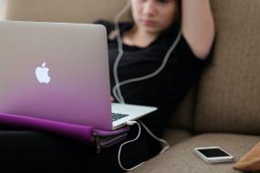 Une jeune fille est assise sur un fauteuil, son ordinateur sur les genoux. Elle a des écouteurs sur les oreilles et un téléphone portable posé à côté d'elle.