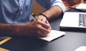 une personne écrit sur une table, un ordinateur portable est placé à sa gauche