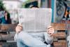 Photographie d'une personne assise sur un banc public tenant un journal. Le journal cache complètement sa tête.